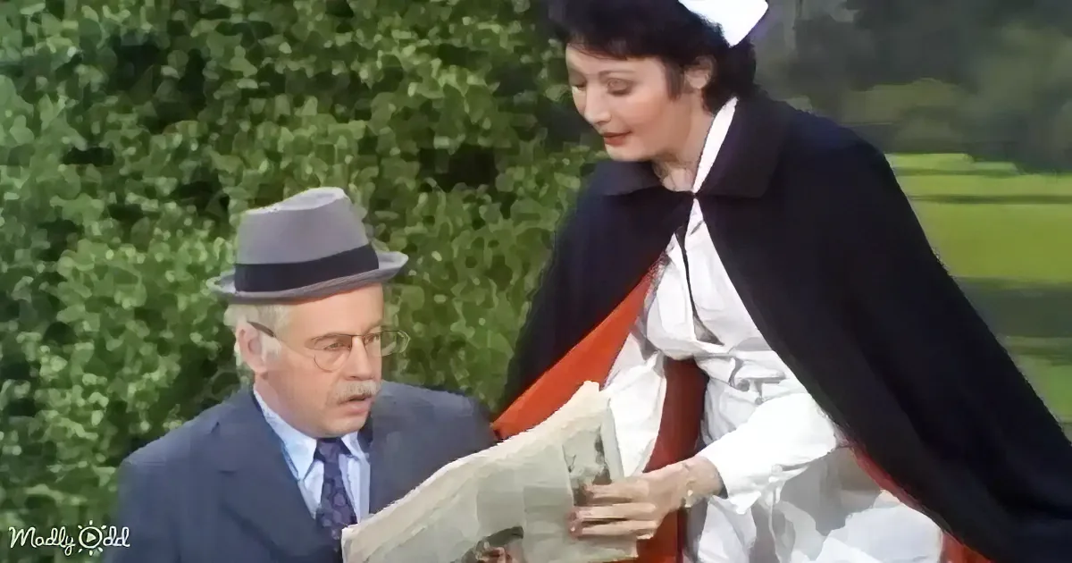 A woman in a black cape and white dress leans over a man in a suit and fedora, who is reading a newspaper.