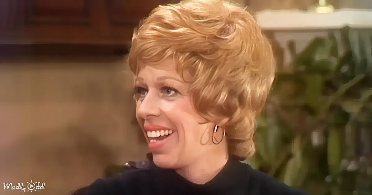 Blonde woman with short hair, wearing a black top, smiling, earrings, indoor setting with plants and furniture.