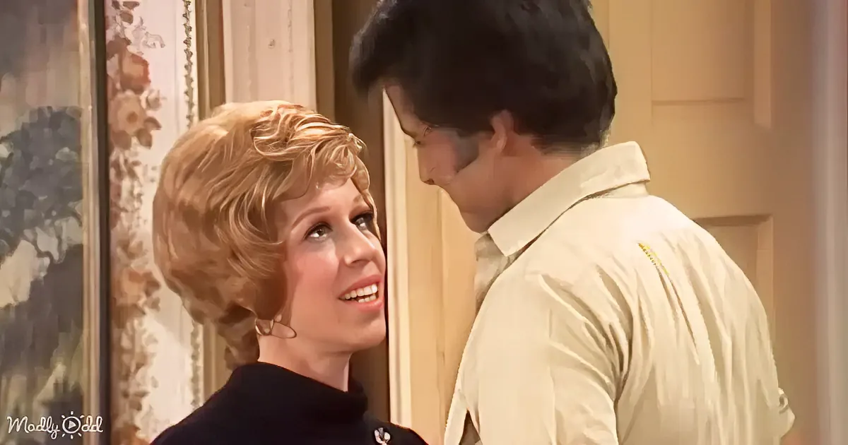 A woman with blonde, curly hair and a man in a light-colored shirt are facing each other in a warmly lit room.