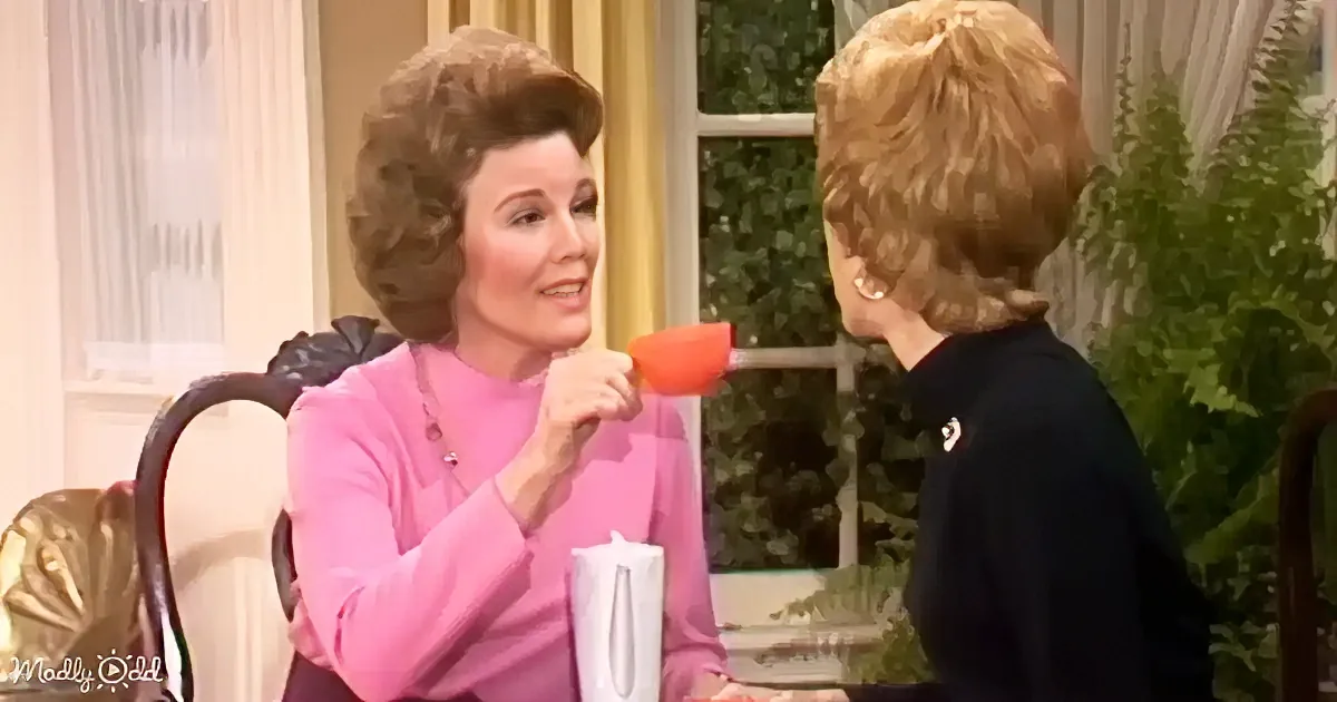 Woman in pink talks into red microphone, seated with another woman in dark top, in a studio setting.