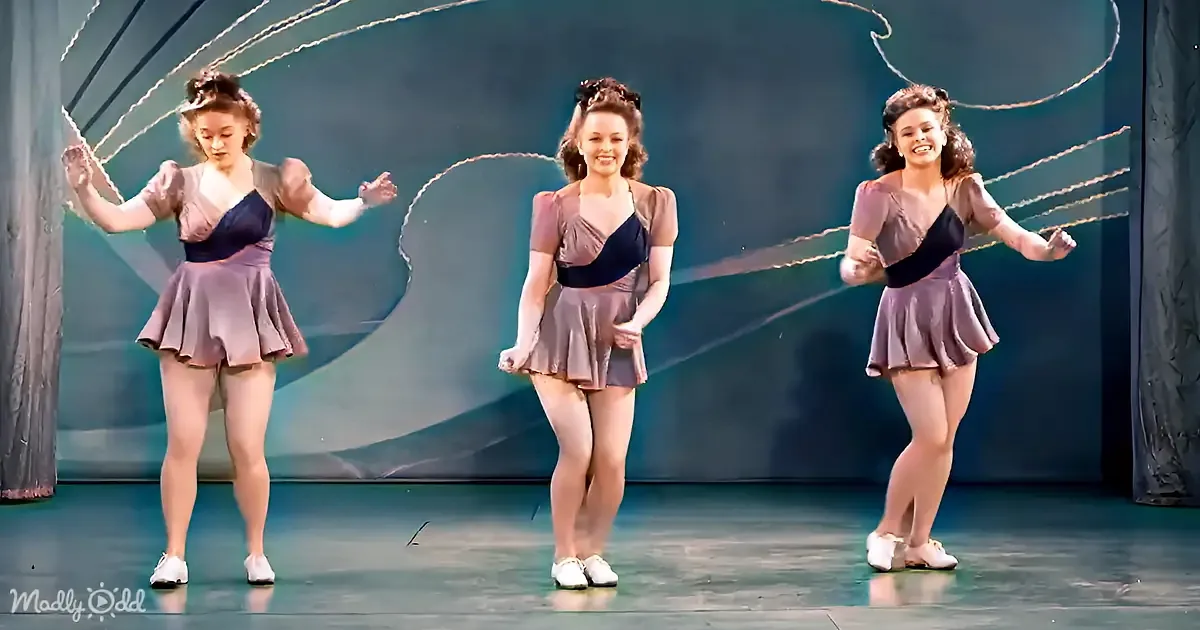 Three women in vintage-style costumes perform a synchronized dance on stage with flowing fabric.