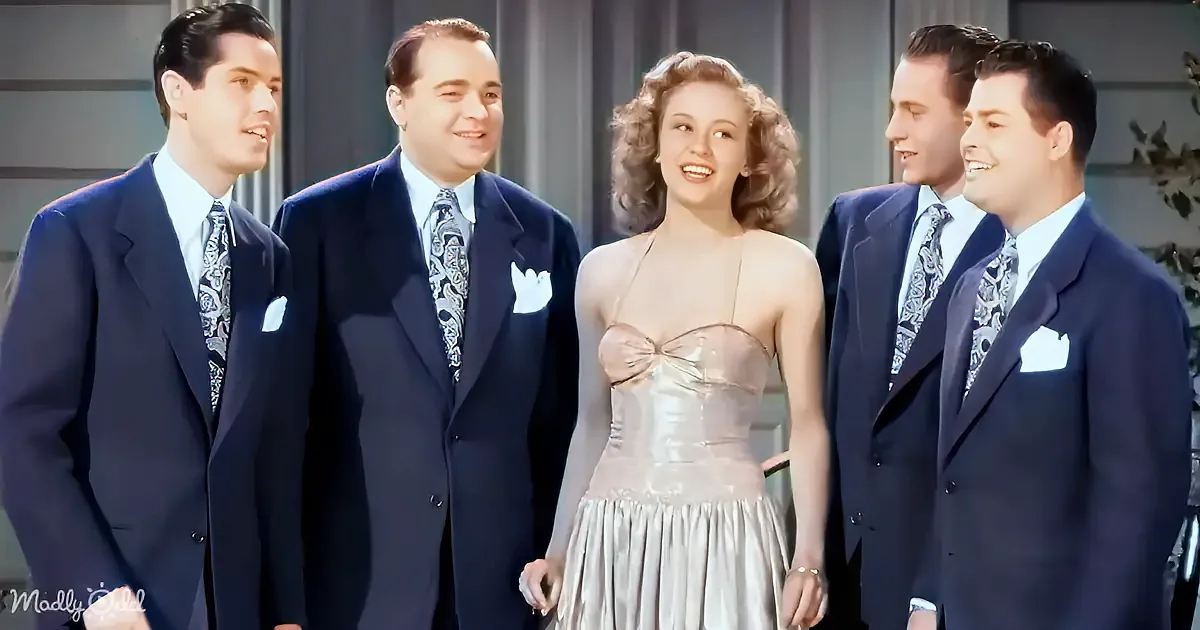 A woman in a silver dress stands with four men in suits, all smiling, in a vintage setting.