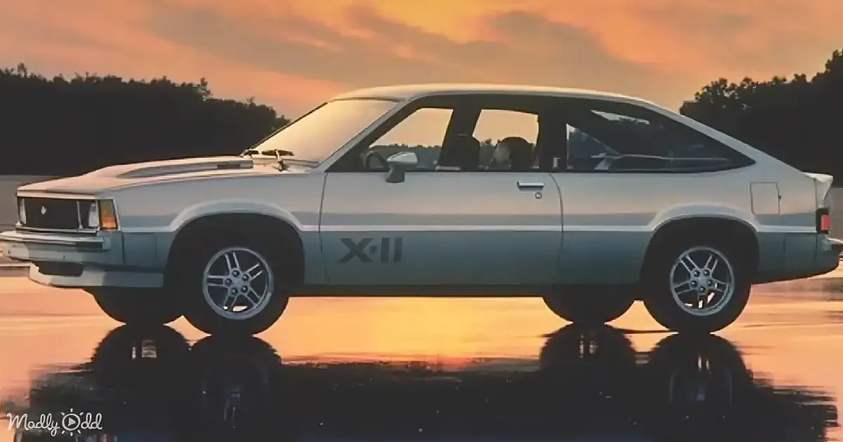 A vintage silver car with "X-11" on its side is parked on a reflective surface at sunset.