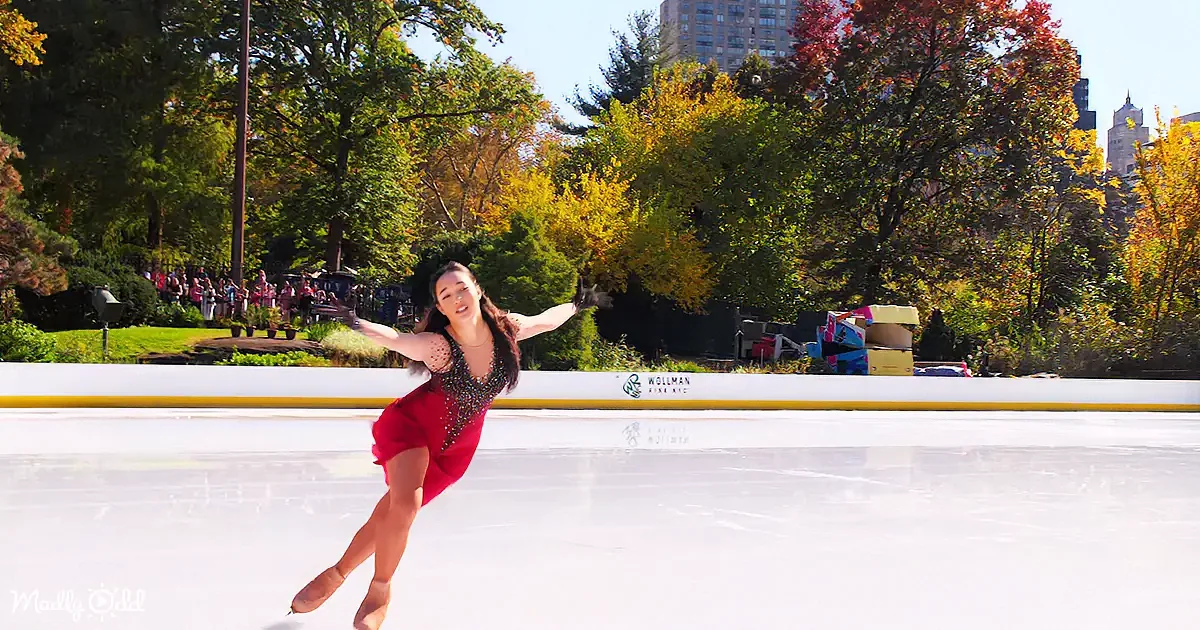 A figure skater in a red dress glides gracefully on an ice rink surrounded by autumn foliage and city buildings.