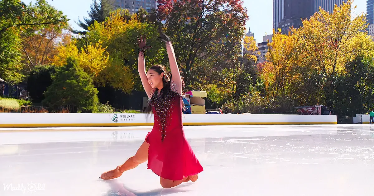 A figure skater in a red dress performs a graceful pose on an ice rink surrounded by autumn trees.