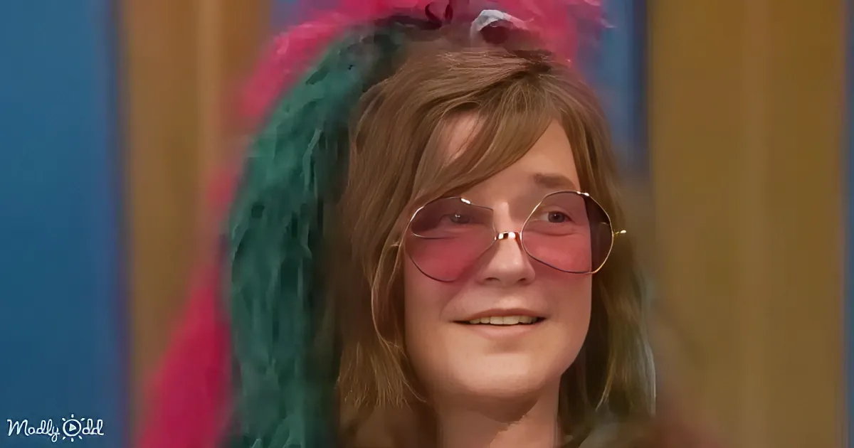 A person with brown hair, wearing pink sunglasses, and a colorful, fluffy headpiece is smiling.