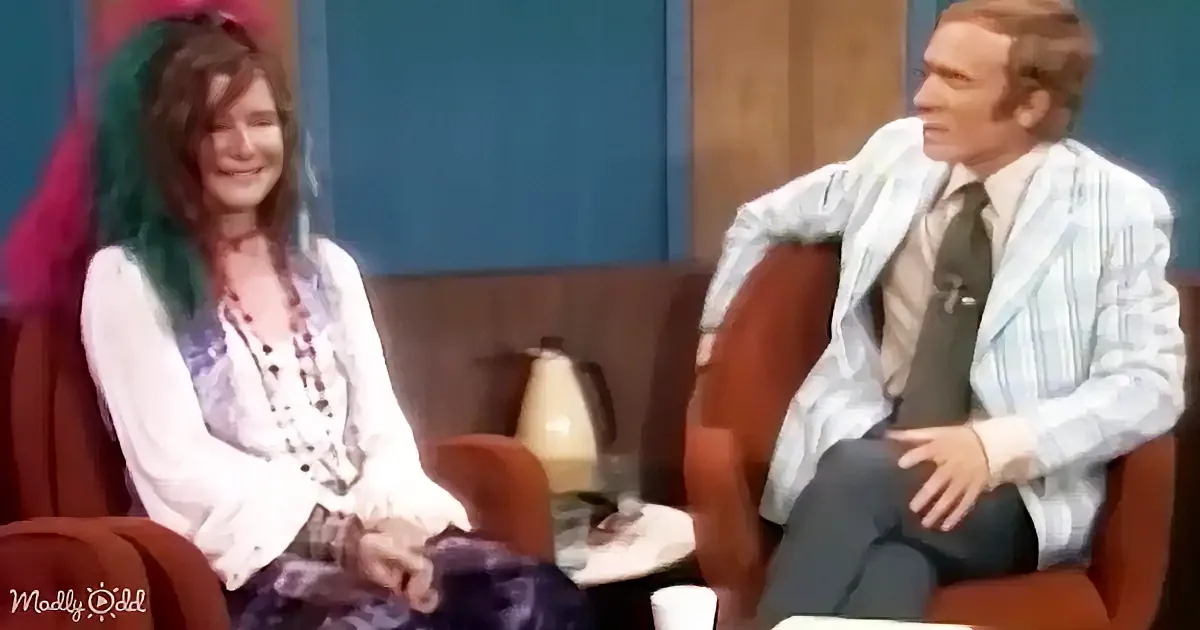 A woman with green hair and a man in a suit sit on a couch, both smiling, in a studio setting.