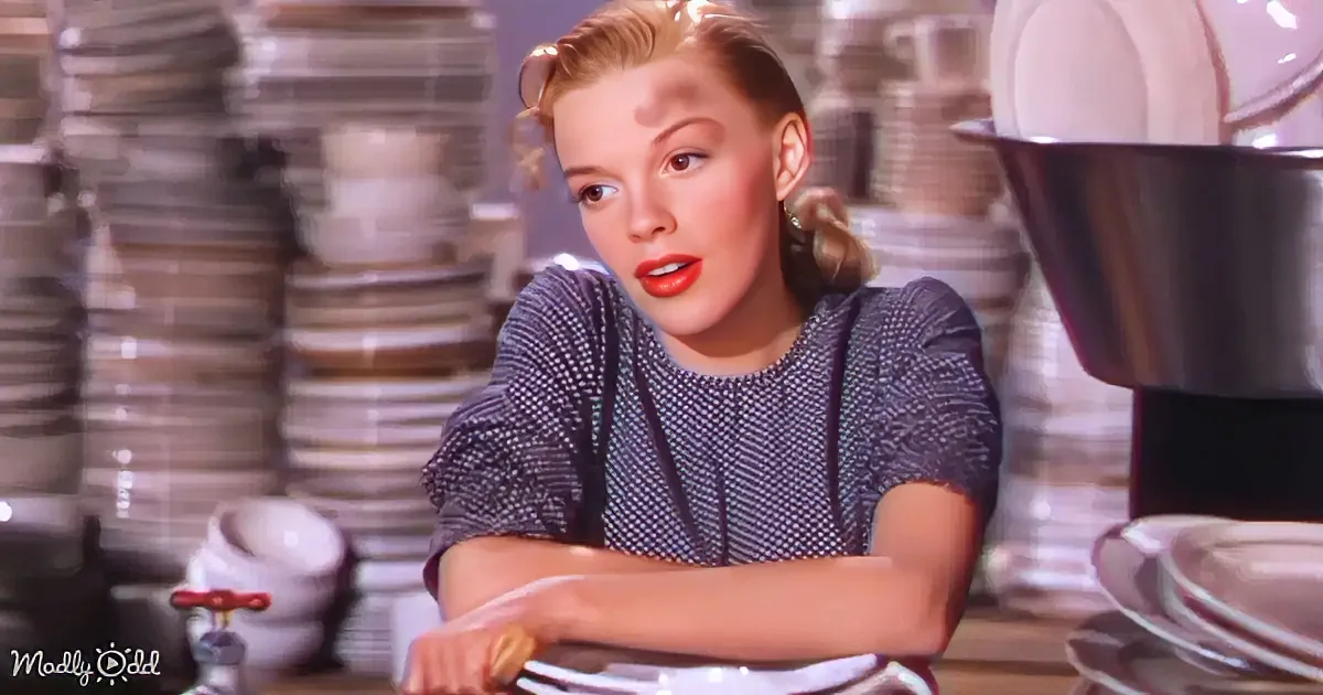 A woman with red lipstick and a patterned top stands in a kitchen with stacks of dishes and a large pot.