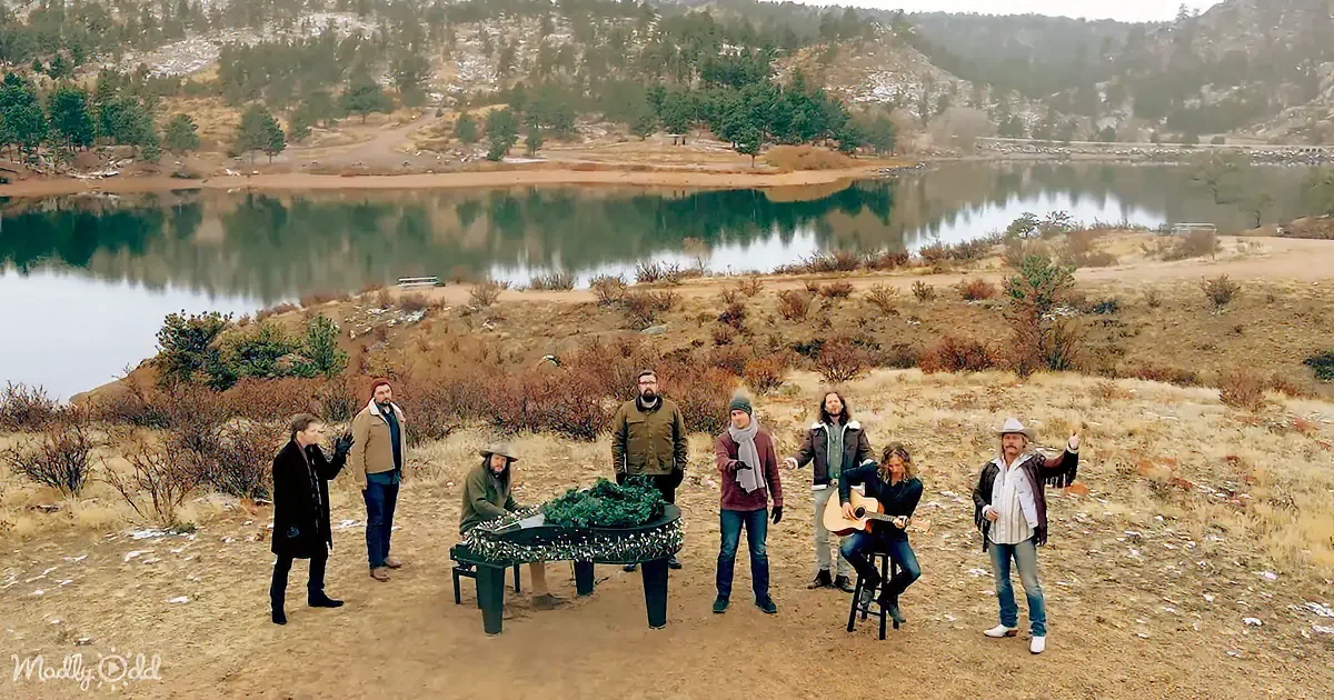 A group of people stands around a table with a Christmas wreath, near a lake with snow-capped hills in the background.