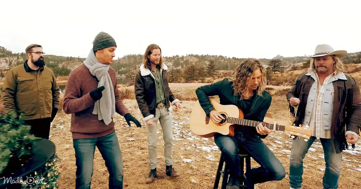 A man in a black jacket plays guitar while others watch in a snowy, rural setting.