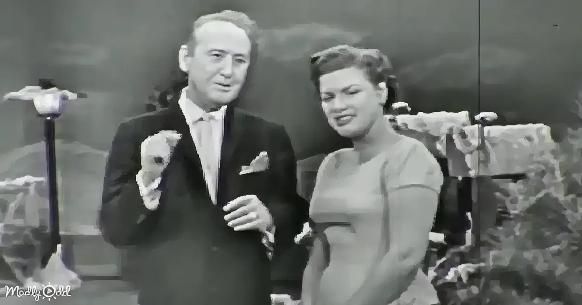 A man in a suit and a woman in a dress stand together, smiling, with a scenic backdrop.