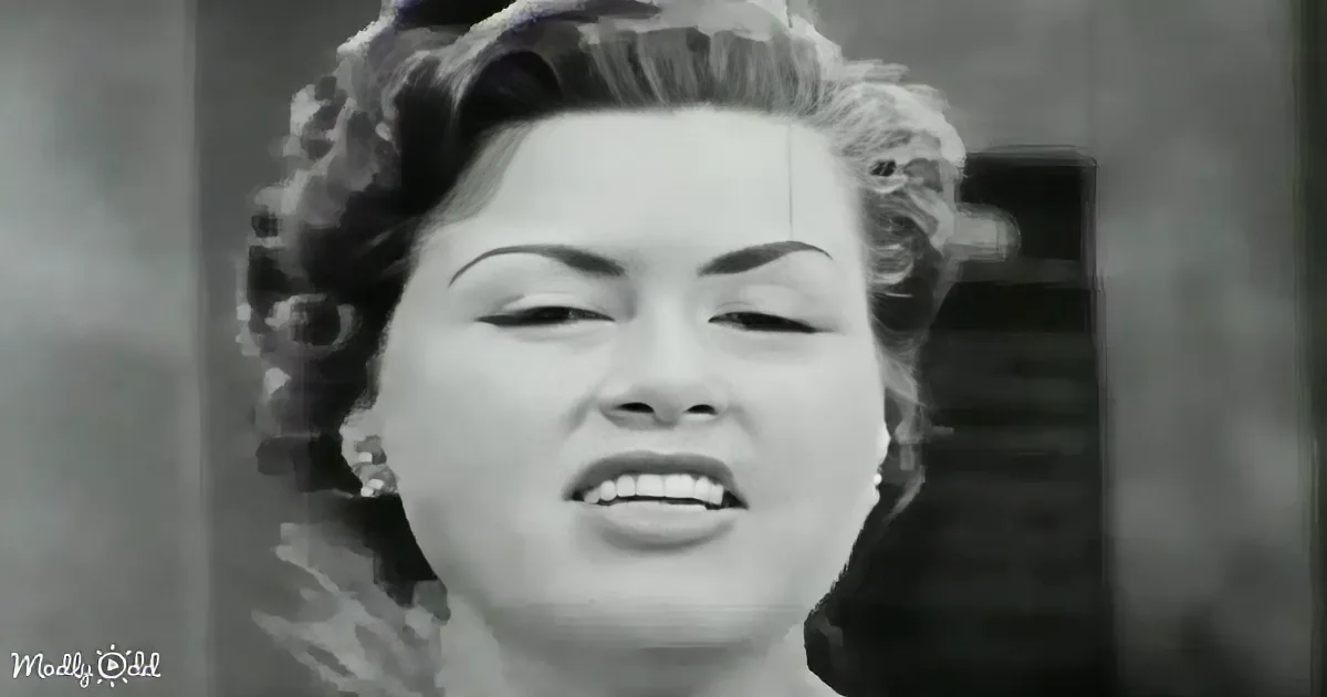 A woman with styled hair and a dark outfit is shown in a close-up, possibly singing or speaking.