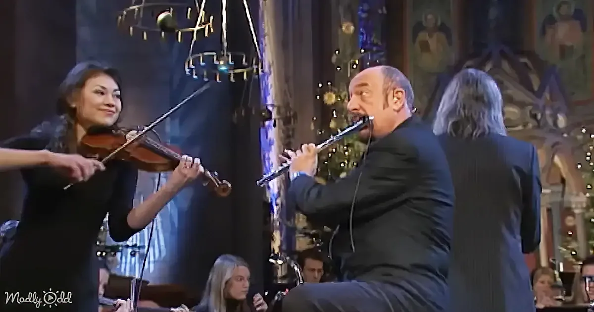 A man in a suit plays a flute while a woman in black plays a violin in front of a decorated Christmas tree.