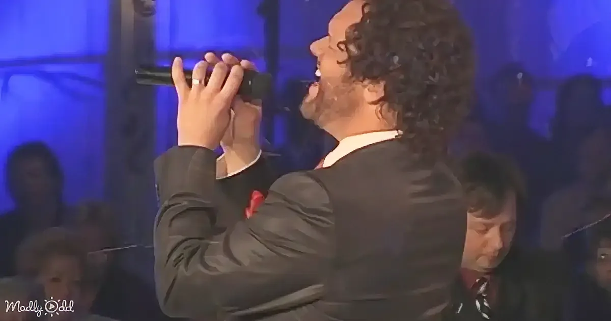 A man in a suit sings passionately into a microphone on stage, surrounded by an audience.
