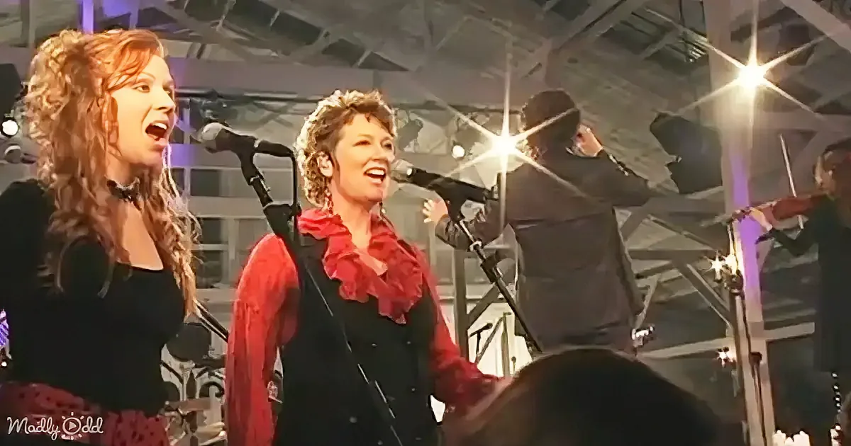 Two women sing on stage, one in red, the other in black, with a band playing in the background.