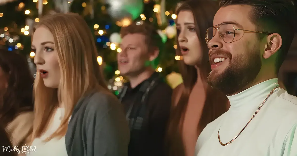 A man with glasses and a beard smiles while singing, surrounded by others in a festive setting with lights.