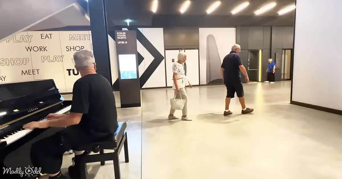 A man plays piano in a modern space with people walking by, walls with "PLAY EAT MEET SHOP" text, and a sign for "Boiler...