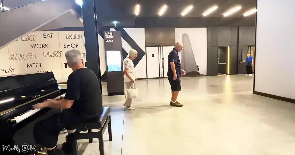 A man plays piano while two others observe in a modern, spacious area with signage and a reflective floor.