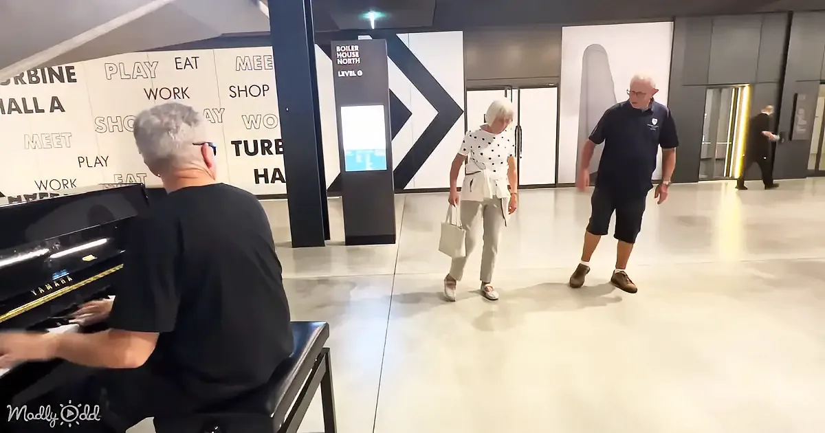 A man plays piano as a woman and man walk by in a modern, open space with directional signage.