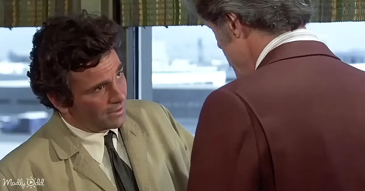 A man in a tan suit and tie is looking at another man in a maroon suit, both in a room with a window view.