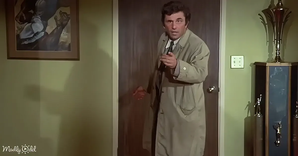A man in a trench coat stands in a doorway, pointing forward, with trophies and a framed picture in the background.