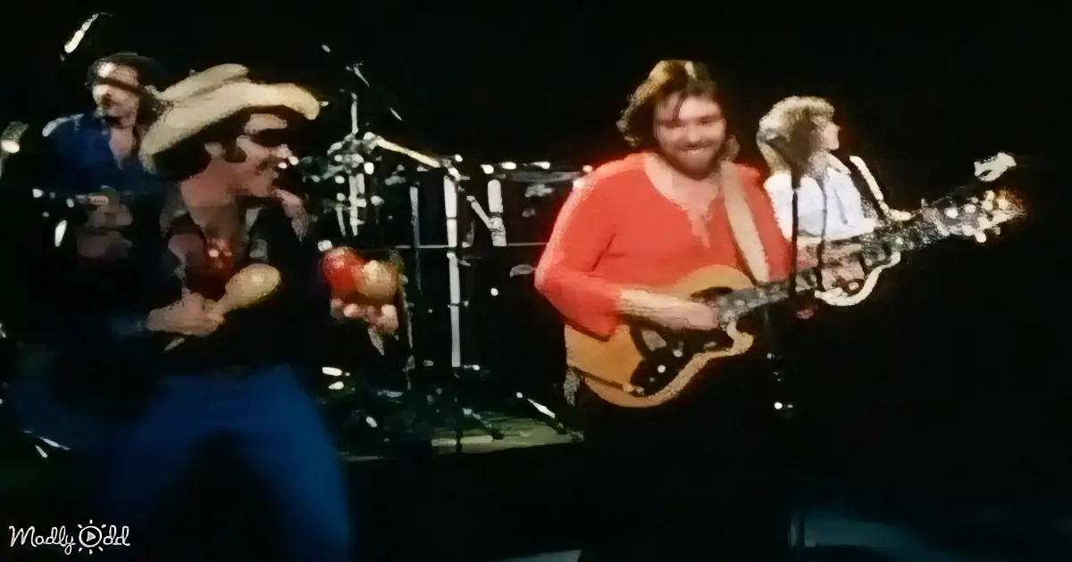 A man in a cowboy hat plays guitar on stage with a band, surrounded by musical instruments and microphones.