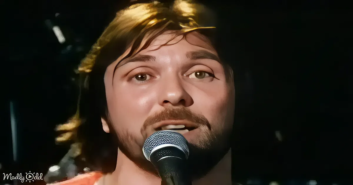 A person with a beard and short hair sings into a microphone, illuminated by a warm light.