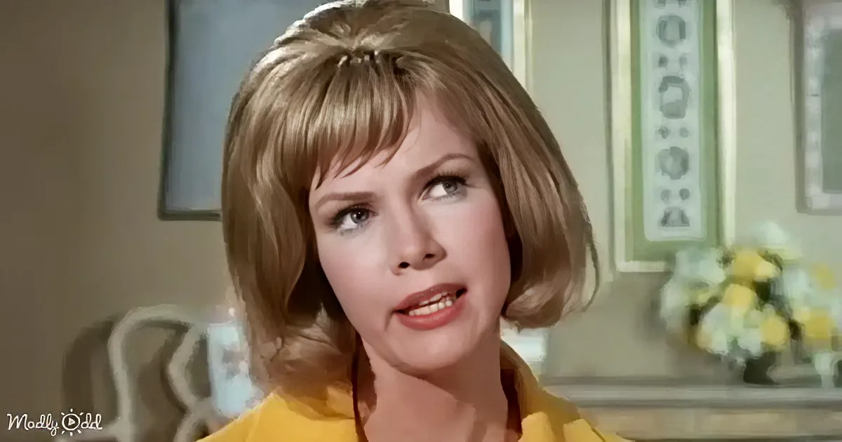 A woman with short blonde hair and a yellow top is looking to the side in a room with framed artwork and flowers.