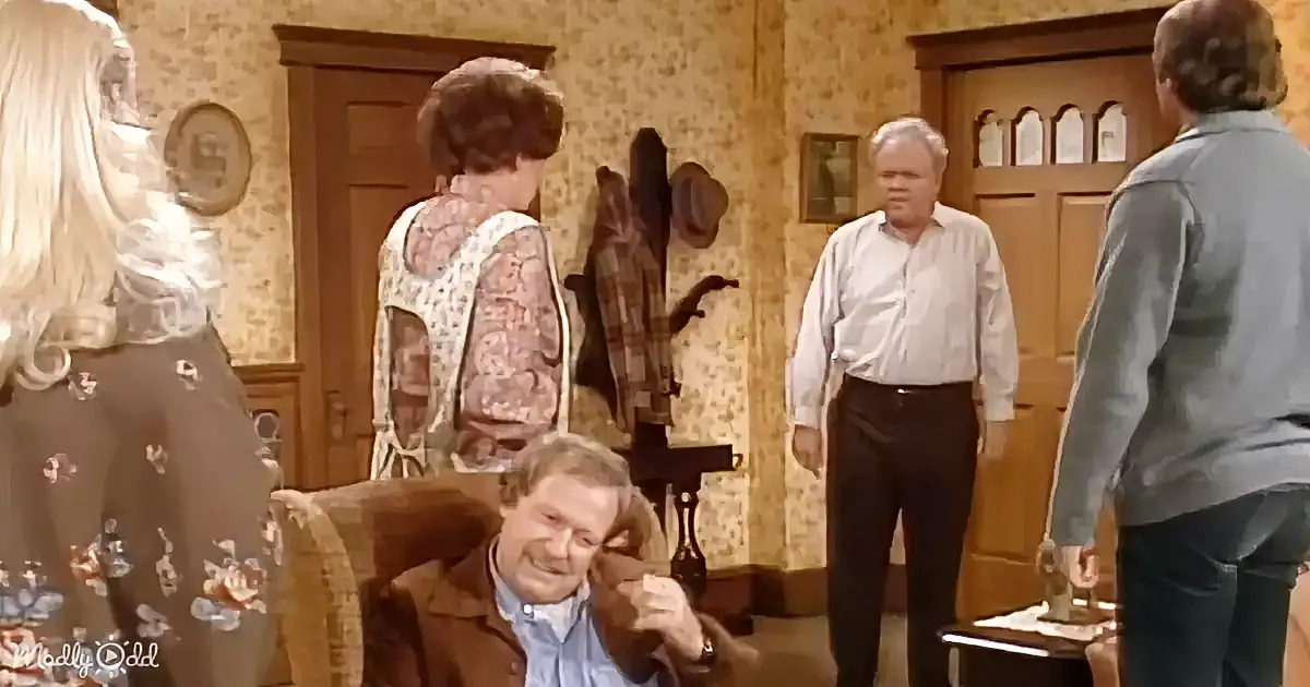 A man in a brown jacket sits in a chair, smiling, while others stand in a room with patterned wallpaper and wooden doors.
