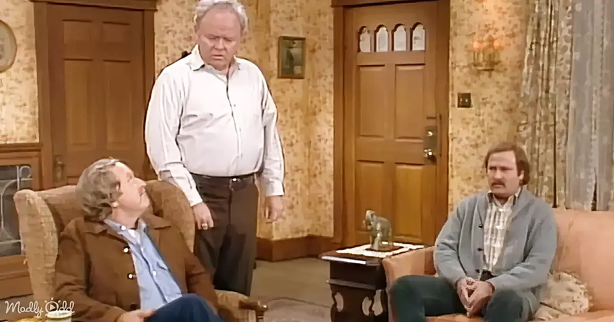 A man in a white shirt stands while another leans back in a chair, and a third sits on a couch in a living room.
