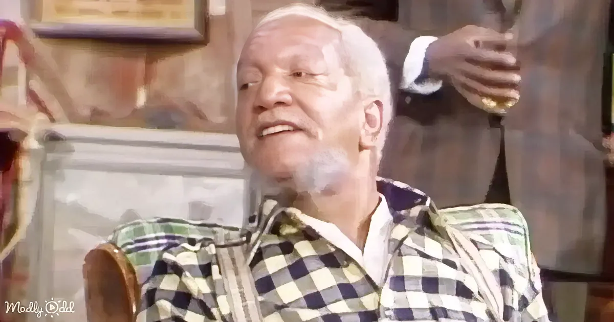 A man in a checkered shirt is sitting, smiling, with a drink in the background.
