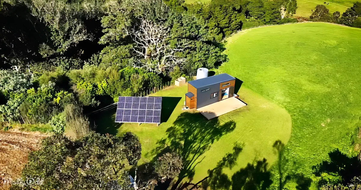 A modern tiny house with solar panels sits on a lush green lawn surrounded by trees.