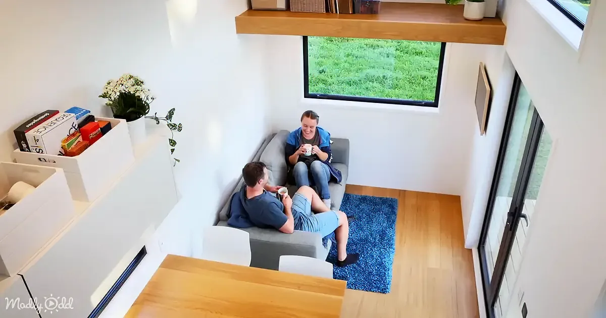 Two people sit on a grey couch in a cozy room with a blue rug, enjoying drinks and a view of green grass outside.