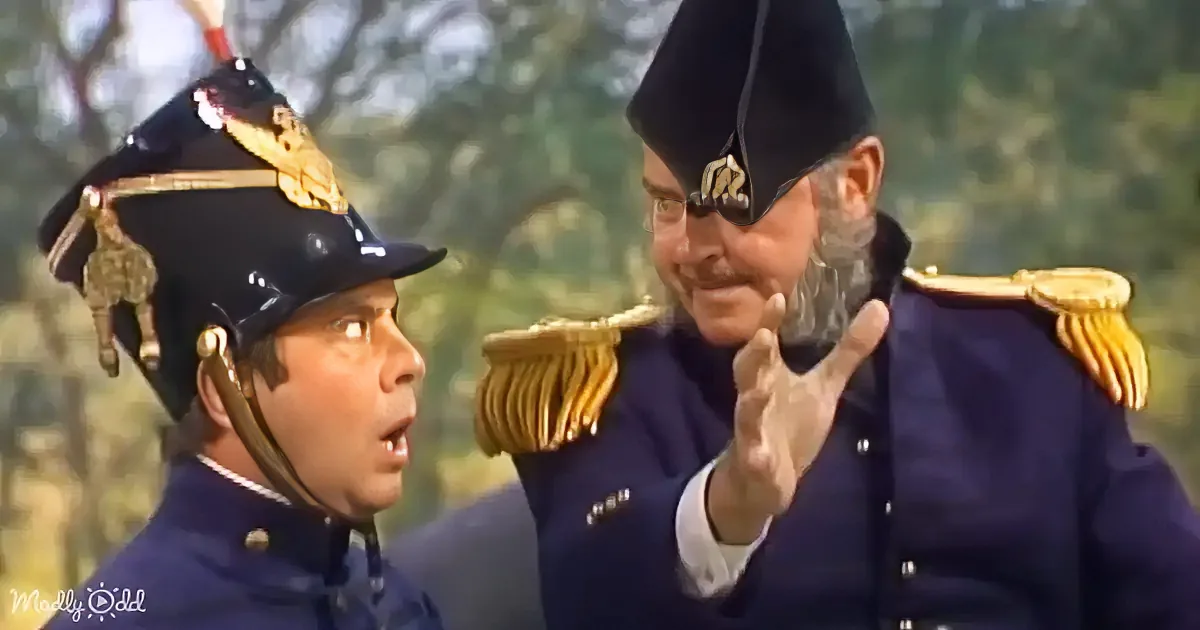 A man in a blue uniform and black hat holds a large black object, possibly a cannon, in a forested area.