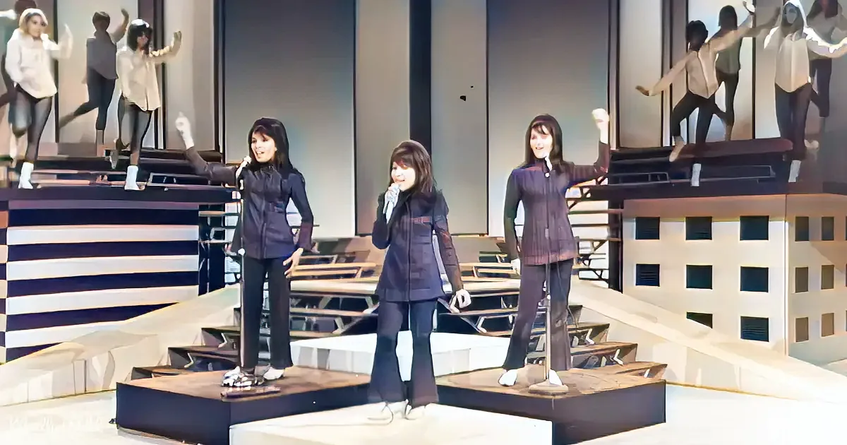 Three women in retro outfits perform on stage with microphones, surrounded by large mirrors.