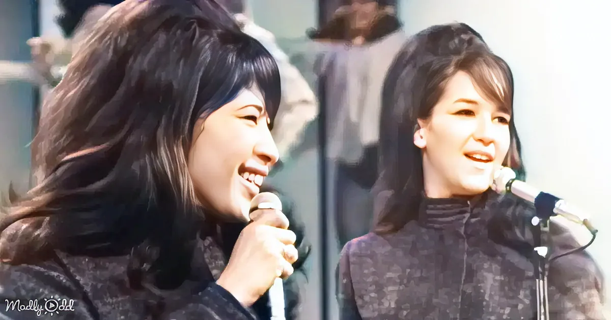 Two women with long dark hair, smiling, holding microphones, standing in front of a window.