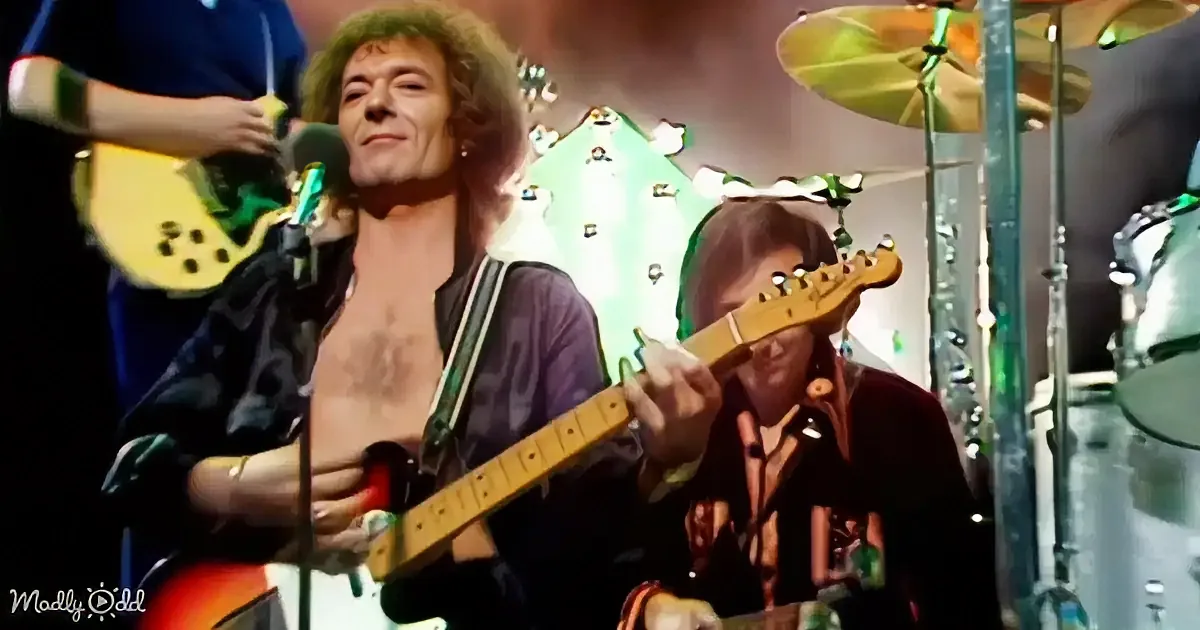 A guitarist with a bare chest plays a guitar on stage, surrounded by other musicians and instruments.