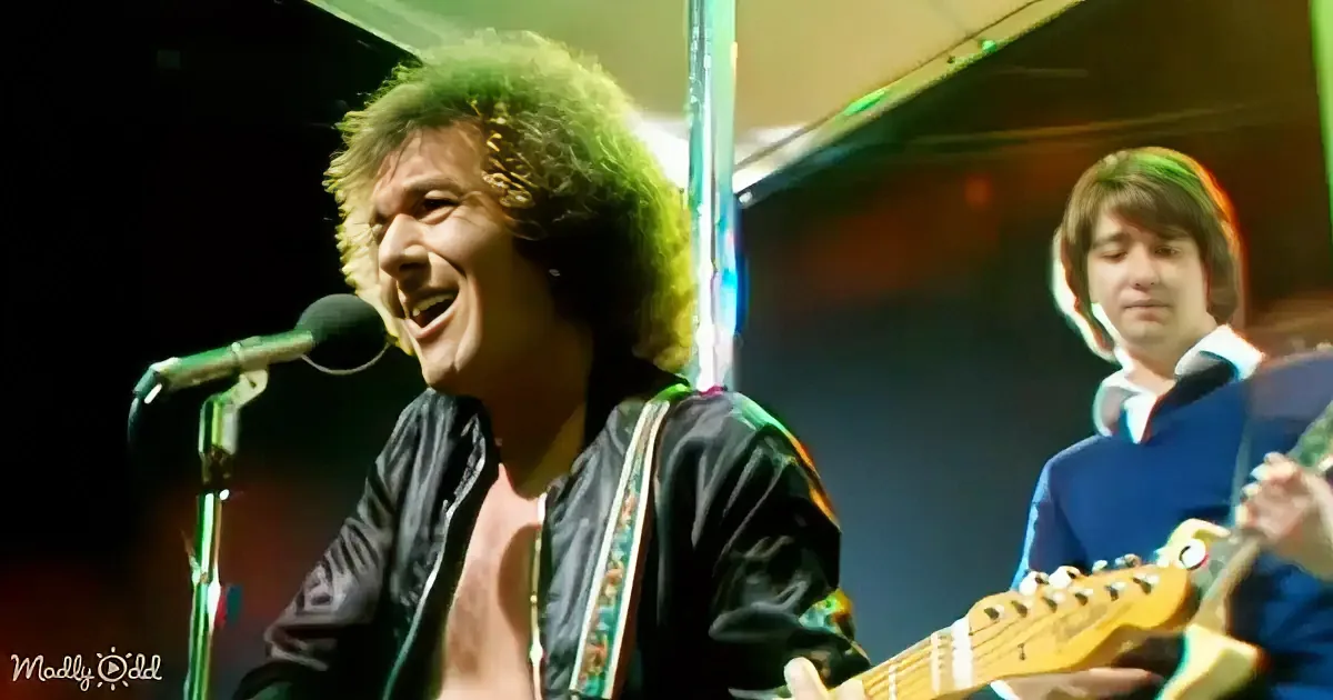 A man with curly hair sings into a microphone while playing guitar on stage.