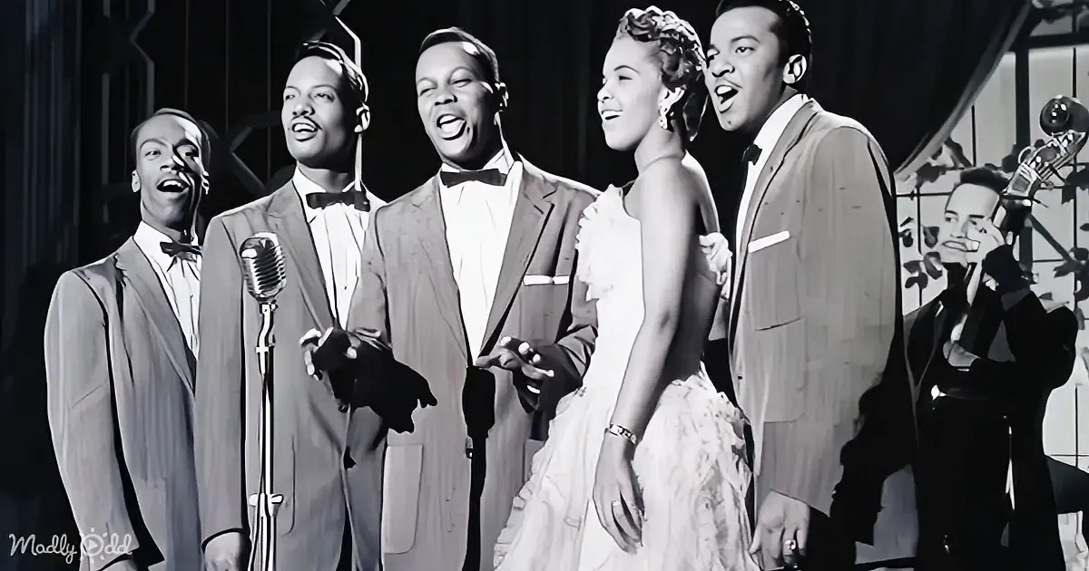 A group of five performers in formal attire sing on stage, accompanied by a musician playing a trumpet.