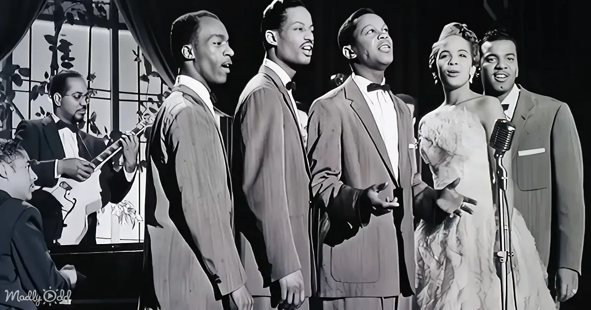 A group of six people, including a woman in a dress, are singing on stage with a musician playing a guitar in the background.