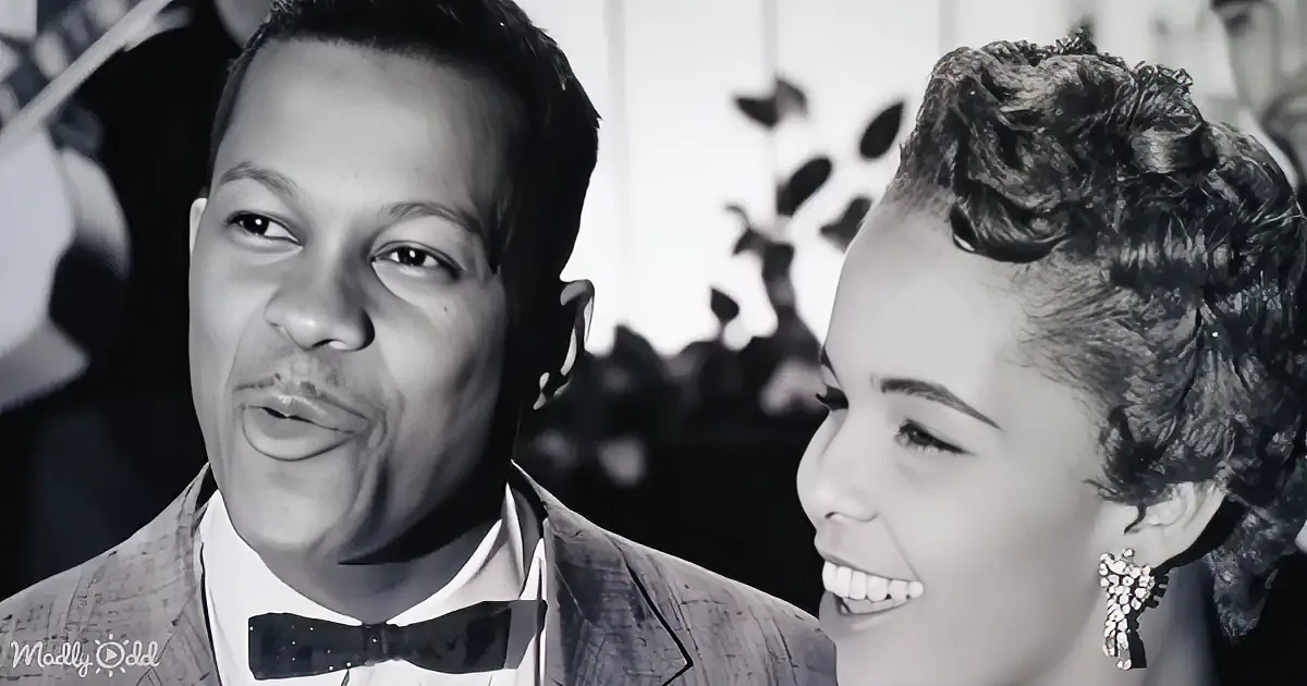 A man in a suit and bow tie smiles alongside a woman with a vintage hairstyle and earrings.
