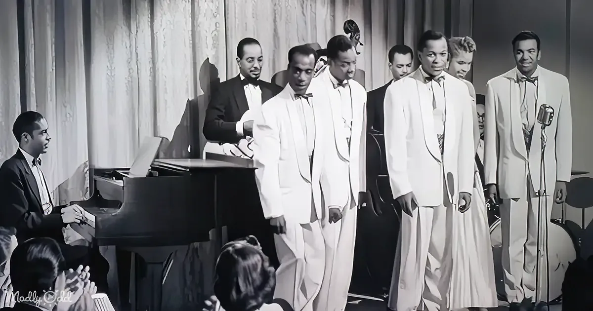 A group of men in white suits sing on stage, accompanied by a pianist, with a curtain backdrop.