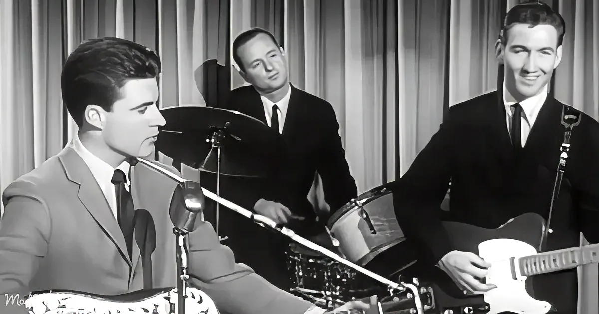 A man in a suit plays guitar while another in a suit plays drums in a vintage musical performance.
