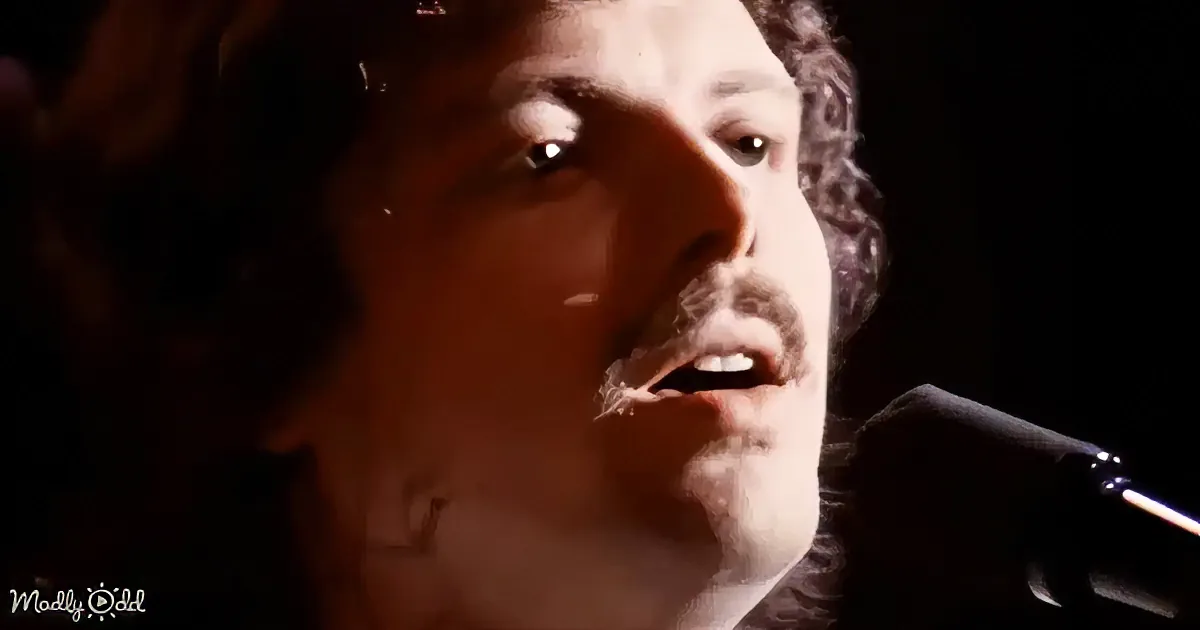 A person with curly hair and a beard is singing into a microphone, with a dark background.