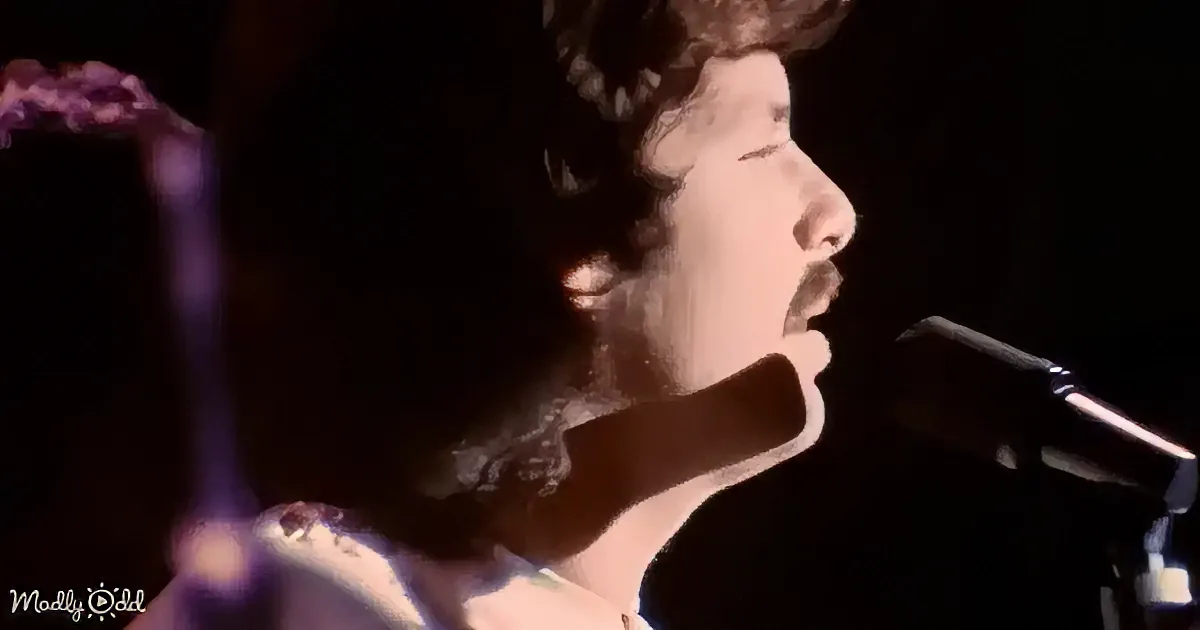 A man with curly hair and a mustache sings into a microphone, illuminated by a spotlight against a dark background.