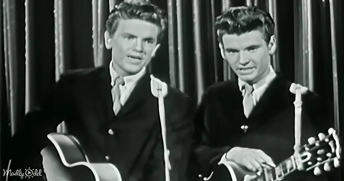 Two men in suits play guitars, one strumming, both seated, against a dark, striped background.