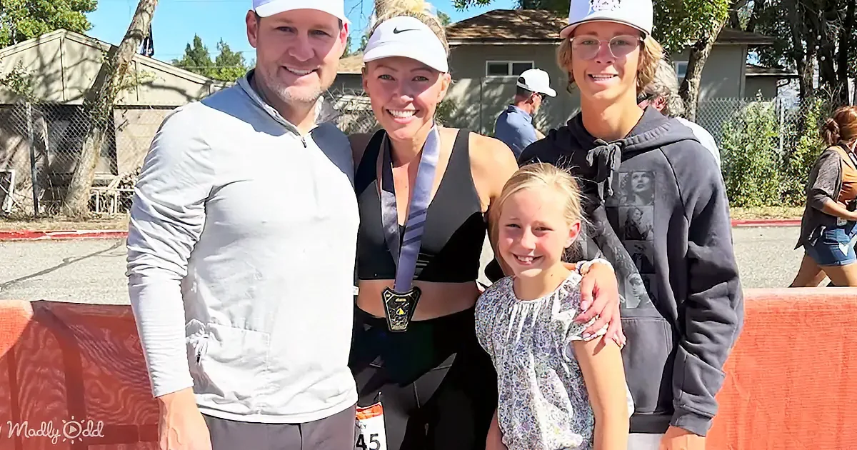 A family of four, including a woman in a sports outfit with a medal, stands together outdoors, smiling.