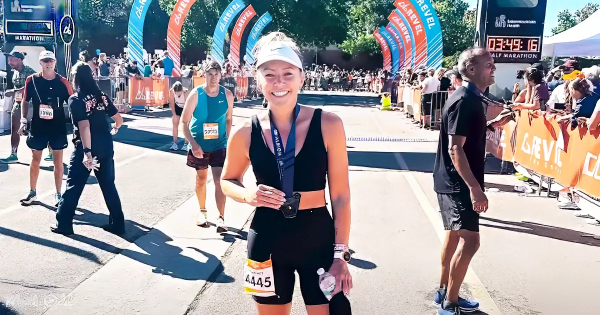 A woman in a black outfit smiles at the finish line of a marathon, holding a medal and a water bottle.