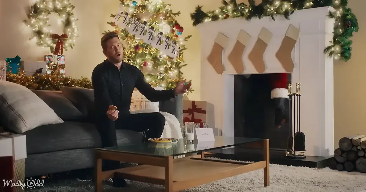 A man sits on a couch in a festive living room, decorated for Christmas, holding a cookie.