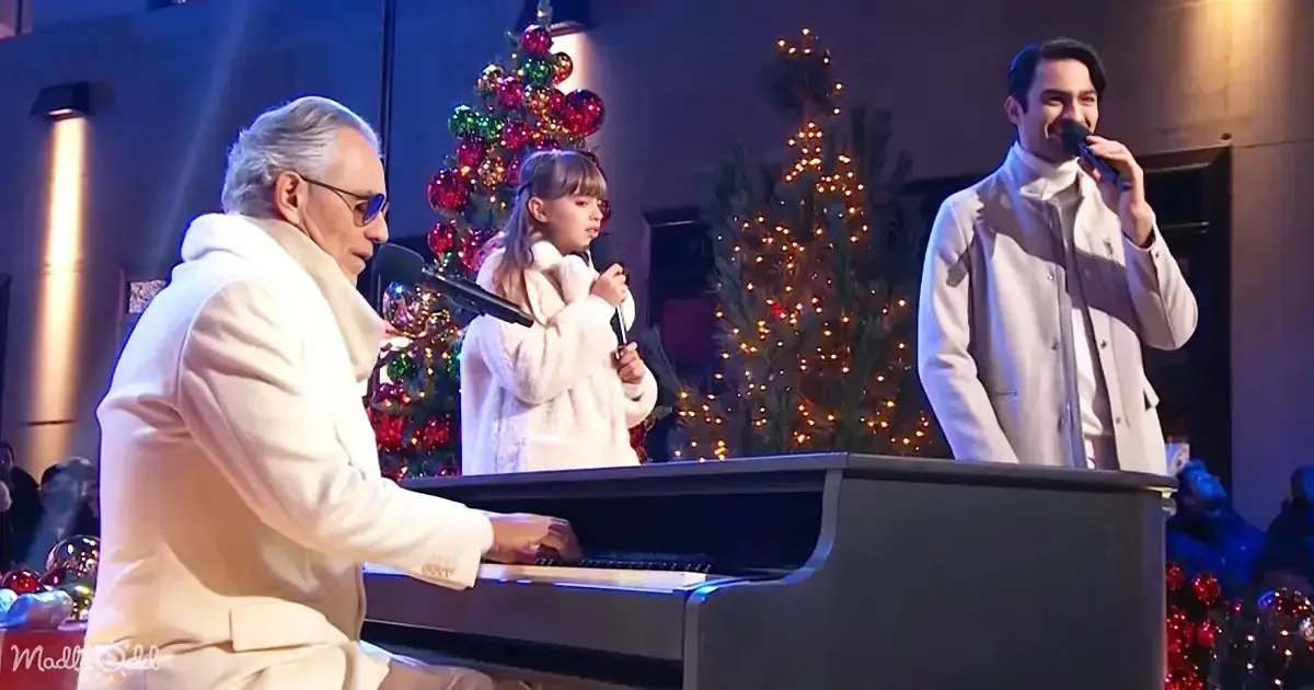 A man in white plays piano while another sings, with a child and Christmas tree in the background.